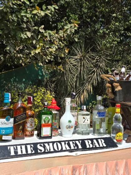 A rustic outdoor bar setup perfect for daytime events. The wooden bar top and backdrop of lush greenery create a relaxed and natural atmosphere for guests to enjoy their drinks.
