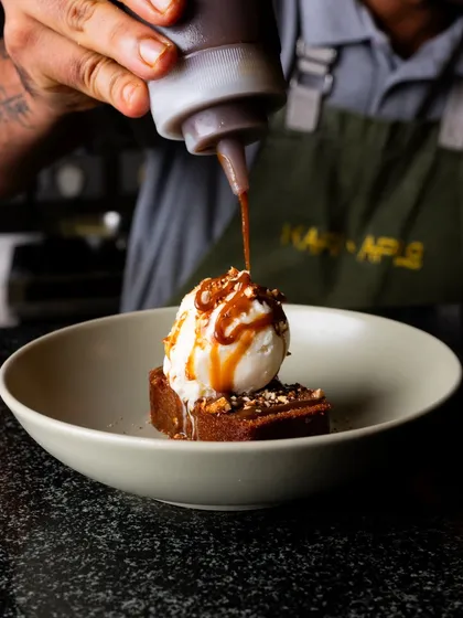 A chef drizzling chocolate sauce over a brownie with ice cream. This action shot captures a moment of creation and indulgence.