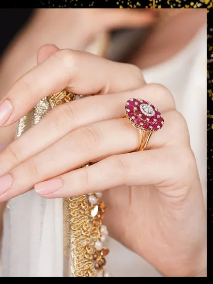 A delicate hand holds a gold-bordered dupatta, drawing attention to a beautiful ruby and diamond floral ring.