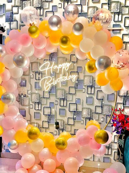 A cheerful and bright balloon ring against a patterned wallpaper. The mix of pink, yellow, white, and silver balloons creates a fun and festive photo corner.