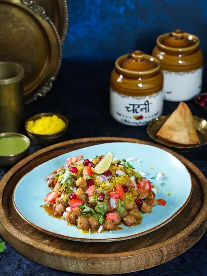 A vibrant bowl of Chana Chaat, garnished with pomegranate, onion, and sev. The dark blue background and traditional chutney jars create a rich, moody setting for this classic Indian snack.
