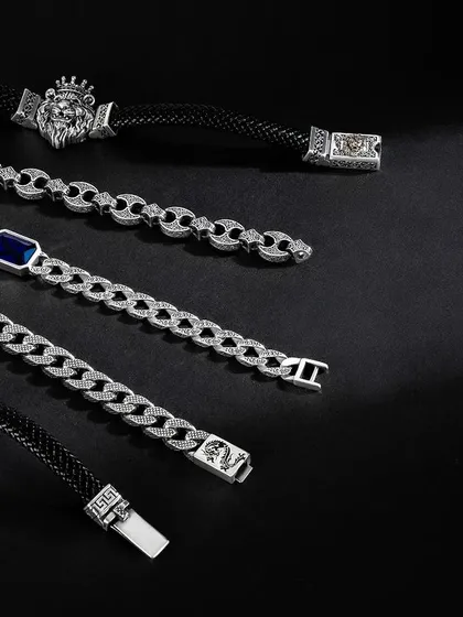 A moody flatlay of men's bracelets and chains. The pieces are arranged diagonally on a black background, with soft lighting that catches the silver and creates a sophisticated, high-end look.