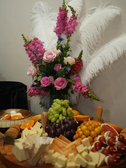 A luxurious spread featuring a dramatic floral and feather arrangement. The board is laden with a variety of cheeses, grapes, and Caprese skewers, perfect for a glamorous celebration.