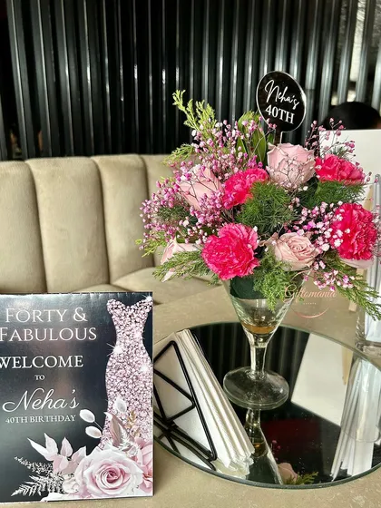 A tabletop decor detail from the 40th birthday party. The custom "Forty & Fabulous" welcome card and a vibrant floral centerpiece in a cocktail glass add a touch of elegance to the guest seating areas.