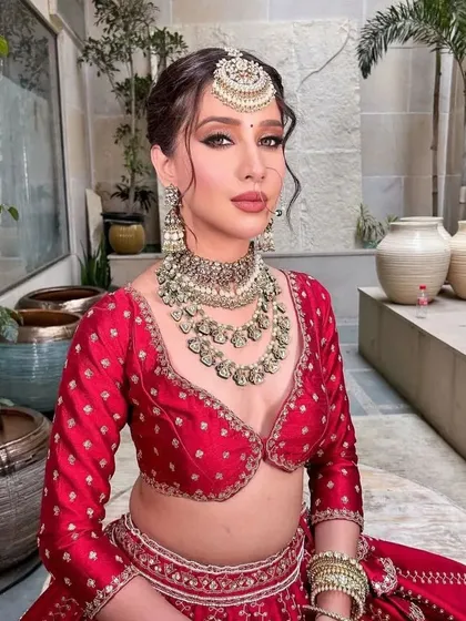 A close-up portrait of the bride. The styling focuses on the intricate maang tikka and layered necklaces that complement the red lehenga.