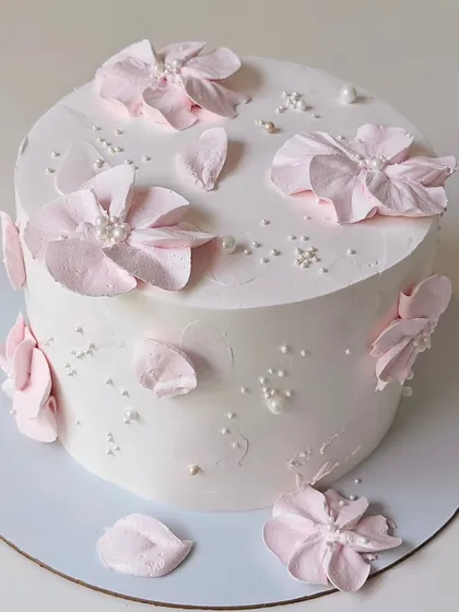 An elegant white cake featuring delicate, hand-sculpted pink wafer paper flowers. The scattered pearls add a touch of sophistication to this minimalist design.