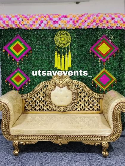 A simple and colorful baby shower backdrop with a green grass wall, a floral top border, and hanging decorative pieces. The golden sofa adds a touch of elegance.