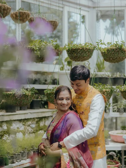 A soft, romantic close-up of the couple in a beautiful greenhouse setting during their Mehendi. The focus is on their happy expressions and gentle embrace, framed by lush greenery.