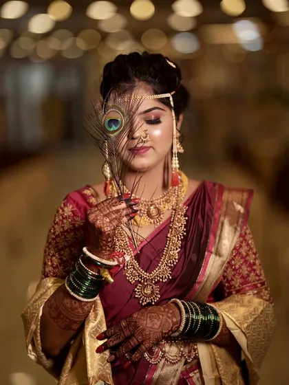 An artistic shot of a Maharashtrian bride. The look features a deep maroon saree, traditional gold jewellery, green glass bangles, and the iconic Mundavalya, with a peacock feather for a touch of grace.