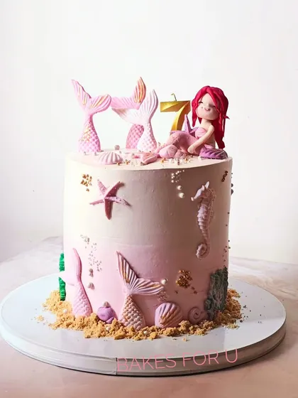 A pretty pink mermaid-themed cake for a 7th birthday. This design features fondant mermaid tails, a cute mermaid figure, and delicate seashell details on a bed of "sand."