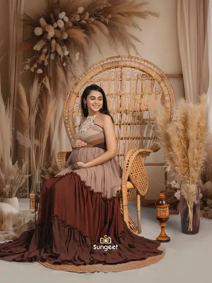A mom-to-be seated in a large peacock chair, part of our boho-chic collection. The tiered brown dress adds to the earthy, natural vibe.