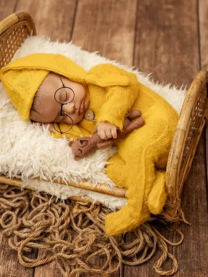 A cozy shot of a baby boy in a yellow sleeper, cuddling a tiny teddy bear on a miniature bed. The round glasses add a touch of whimsy.