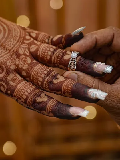 A close-up of the ring ceremony, highlighting the bride's detailed mehendi. The design features the engagement ring symbol on the palm, which mirrors the actual ring being placed on her finger.