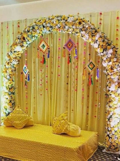 A simple yet elegant Haldi seating arrangement. The backdrop is a floral arch made of yellow and white flowers, with hanging tassels for a pop of color.
