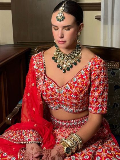 A serene bride captured in a moment of reflection. Her makeup is soft yet defined, with a sleek bun hairstyle that beautifully showcases her emerald green necklace and maang tikka.