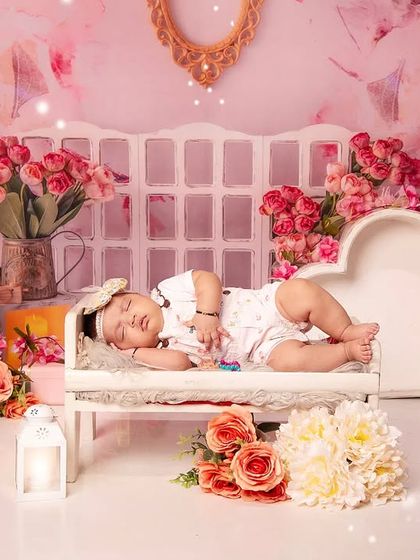 This baby is sleeping on a miniature white chaise lounge, set against a soft pink floral backdrop. The entire scene is filled with beautiful bouquets, creating a truly romantic portrait.