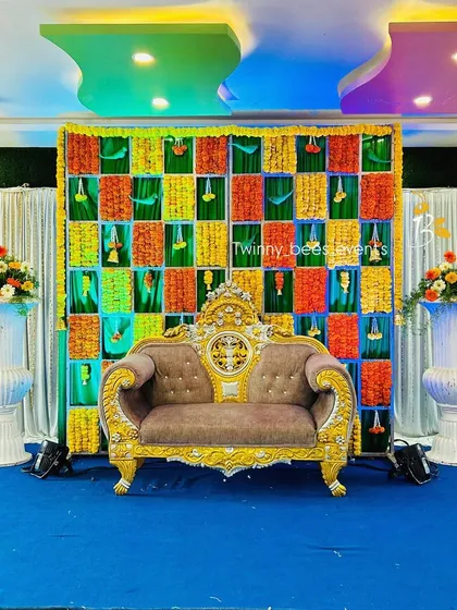 A creative and colorful checkerboard backdrop made from panels of yellow marigolds and orange and green fabric. This unique design is perfect for adding a festive and modern touch to events like baby showers or naming ceremonies.