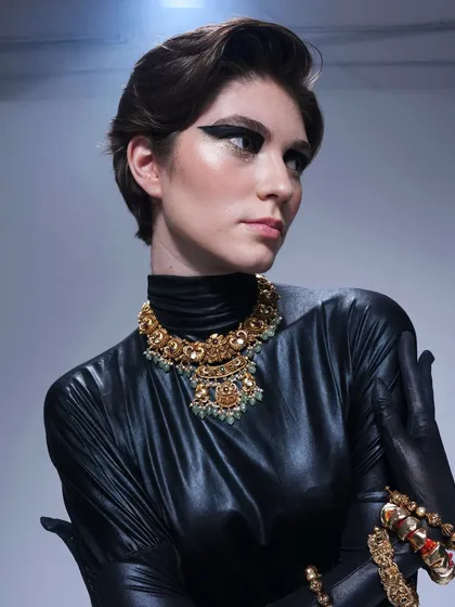 A portrait from the same high-fashion shoot, showing the contrast between the sleek black outfit, the traditional gold jewelry, and the modern, graphic eye makeup.
