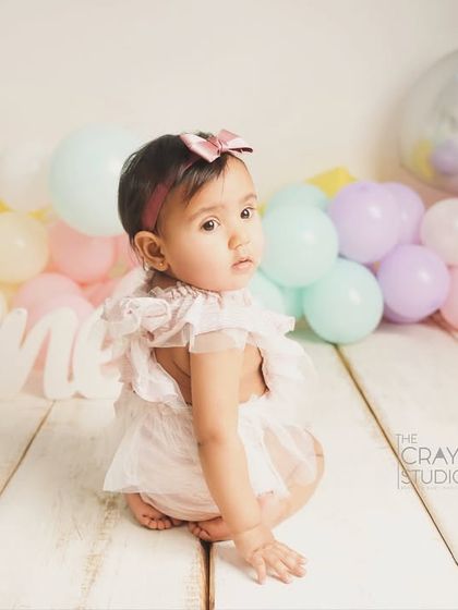 A sweet, shy look over the shoulder from the birthday girl, sitting amongst pastel balloons and a "One" sign.