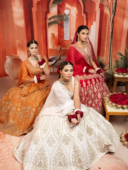 A moment of regal poise. This image showcases three distinct bridal lehengas in shades of red, orange, and ivory, each telling its own story through unique embroidery and silhouette.
