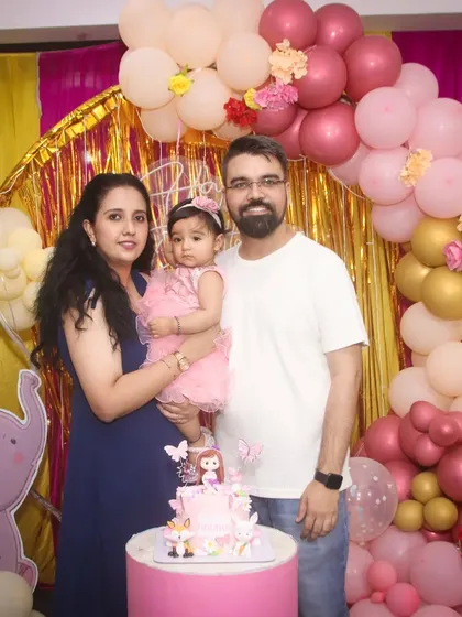 A classic family photo from a first birthday party. The family poses together with the cake against a beautiful pink and gold balloon arch, creating a perfect memory of the celebration.