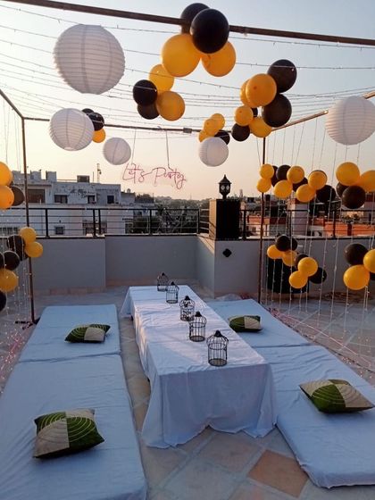 A daytime view of a terrace party setup. The space is decorated with yellow and black balloons, white paper lanterns, and a low seating arrangement for a casual celebration.