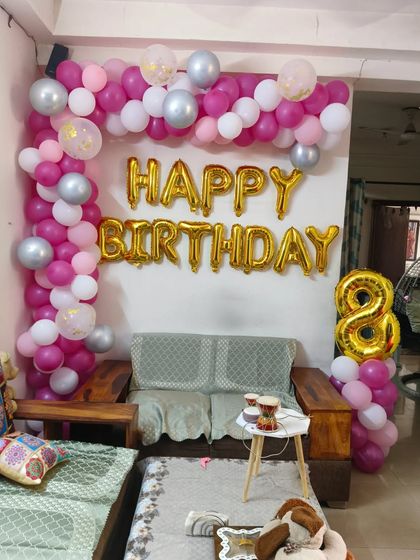 A simple and sweet 8th birthday setup at home. A pink, white, and silver balloon arch frames the sofa, complemented by 'Happy Birthday' and number '8' foil balloons.