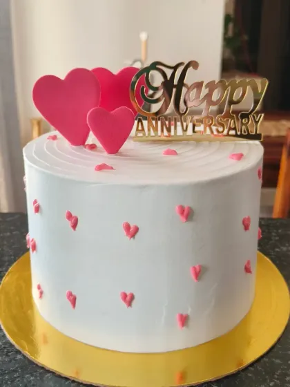 A classic white anniversary cake dotted with little pink hearts and topped with a gold acrylic topper.