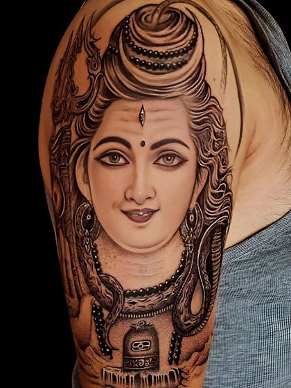 A serene portrait of Lord Shiva on the upper arm, detailed with a Shivling and cobra. This black and grey tattoo focuses on a calm and meditative expression.