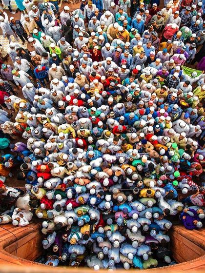 A unique, tightly framed overhead shot looking down into one of the mosque's towers, with a dense crowd of worshippers visible below. The colorful clothing and white prayer caps create a vibrant mosaic of faith.