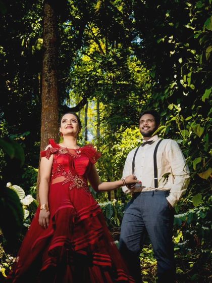 A creative pre-wedding photo taken in a lush, green forest. The couple's outfits, a red gown with illusion details and a vintage-style shirt with suspenders, create a unique look.