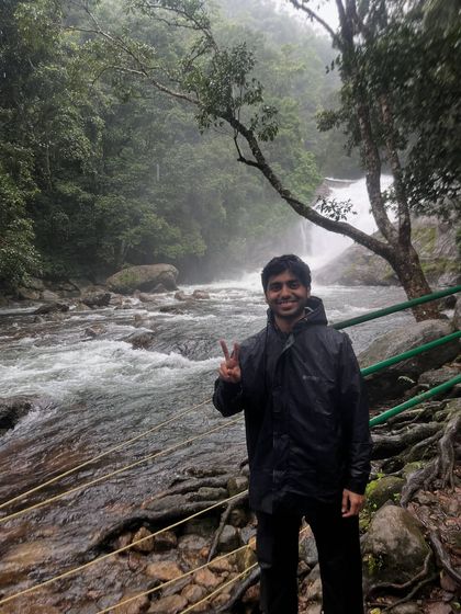 A trekker gives a peace sign while standing by a gushing river and waterfall in Munnar.