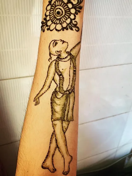 A clear, still image of the Gaura Hari devotional henna. This design covers a large part of the forearm, with a delicate floral mandala pattern above the main figure.