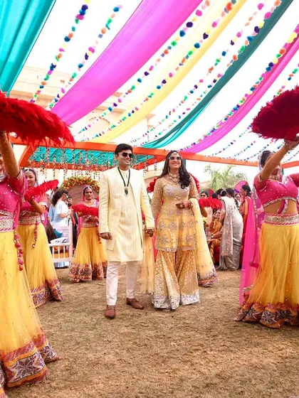 The couple's grand entry at their Mehendi carnival, surrounded by dancers with red feathered fans, creating a visually stunning moment.