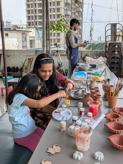 This wide shot shows a mother and daughter painting at a long table filled with pottery, showcasing the communal and creative atmosphere of a family session.