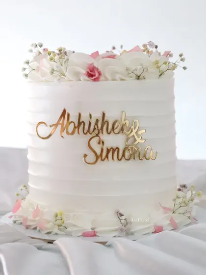 A beautiful single-tier cake for an engagement or anniversary. It features a clean, textured buttercream finish, a custom gold name topper, and is crowned with fresh flowers.