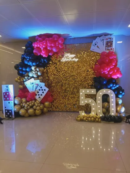A fun casino-themed 50th birthday party. The centerpiece is a gold sequin wall, decorated with playing card props, marquee numbers, and a balloon arch in red, black, and gold.
