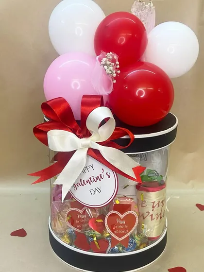 A fun and festive Valentine's hamper in a clear cylindrical box, filled with chocolates and treats and topped with a red, white, and pink balloon bouquet.