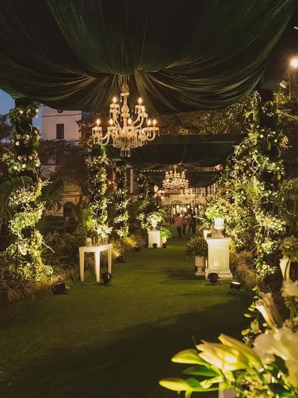 A magical entrance walkway for an evening wedding. The path is lined with lush greenery and white flowers, with elegant chandeliers and green velvet drapes overhead creating a dramatic and enchanting effect.
