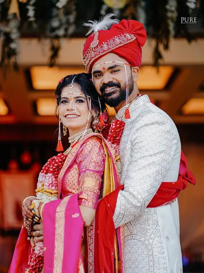 A classic portrait of a smiling Marathi couple on their wedding day. The bride's pink and yellow saree contrasts beautifully with the groom's white sherwani.