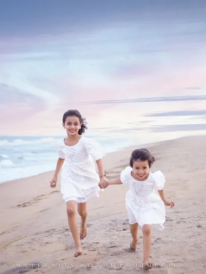 Two sisters holding hands and running freely on the sand. This sibling photograph captures the carefree spirit of childhood against the beautiful backdrop of the sea and a soft, painted sky.