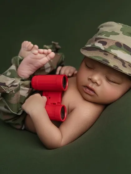 This little soldier is sleeping soundly, dressed in camouflage pants and a cap while holding a pair of bright red binoculars. It's a wonderful tribute for families with a military background.