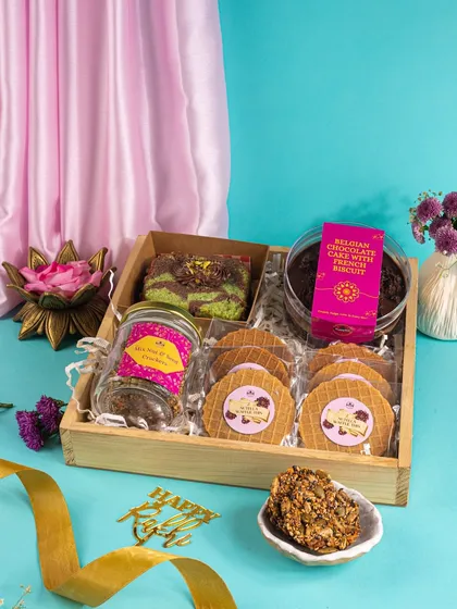 This Rakhi hamper is styled in a wooden tray against a soft blue and pink backdrop. The composition highlights the variety of treats, from waffles and cookies to a jar of cake.