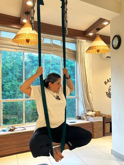 A student holds a supported squat in the aerial hammock, a pose that builds lower body strength and stability. The natural light from the large windows adds to the serene atmosphere of the studio.