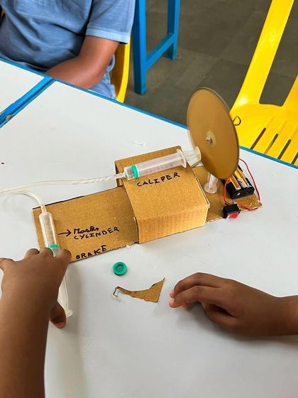 A student proudly displays their completed disc brake model, built from cardboard and syringes to demonstrate the principles of hydraulics.