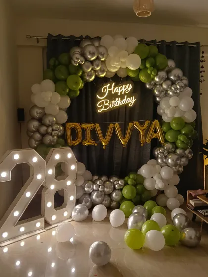 A 28th birthday party with a circular balloon arch in green, silver, and white, complemented by a neon sign and large marquee numbers.