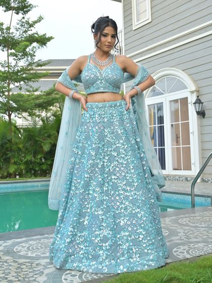 A full-length shot of a model in a sequined blue lehenga by the pool. The halter neck blouse is paired with a matching diamond necklace and bracelets for a chic, modern look.