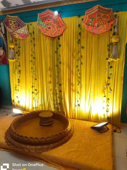 A simple and traditional Haldi backdrop for a home function. The setup includes a bright yellow curtain, decorative umbrellas, and a low wooden stool (chowki) for the ceremony.