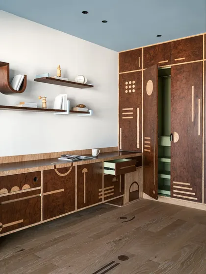 Storage should never be boring. For this multipurpose room, we designed a full-wall unit in rich burl veneer with intricate geometric inlays. The design combines different shutter types and playful motifs, with a surprise pop of mint green inside, turning a practical piece into a visual delight.