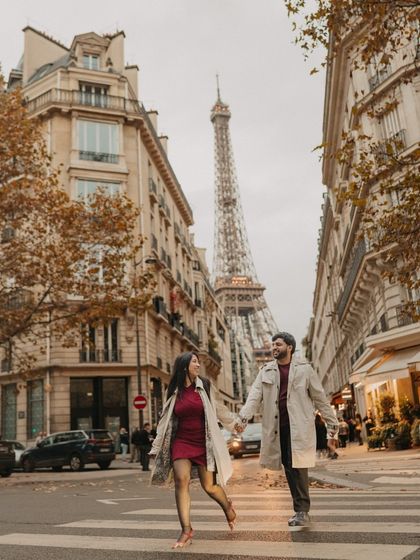 A classic "crossing the street" photo in Paris, with the Eiffel Tower in the background. The couple's coordinated trench coats and joyful expressions make for a stylish and timeless image.
